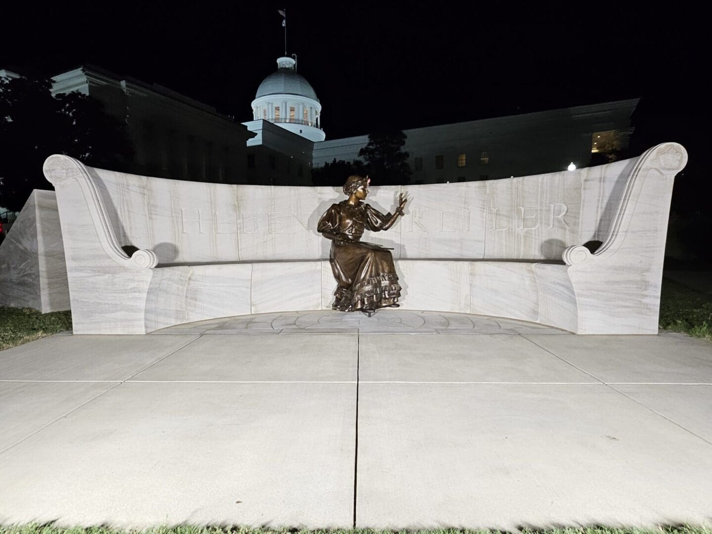 Curved white marble memorial with a bronze figure at its centre, photographed at night with a capitol dome lit behind it