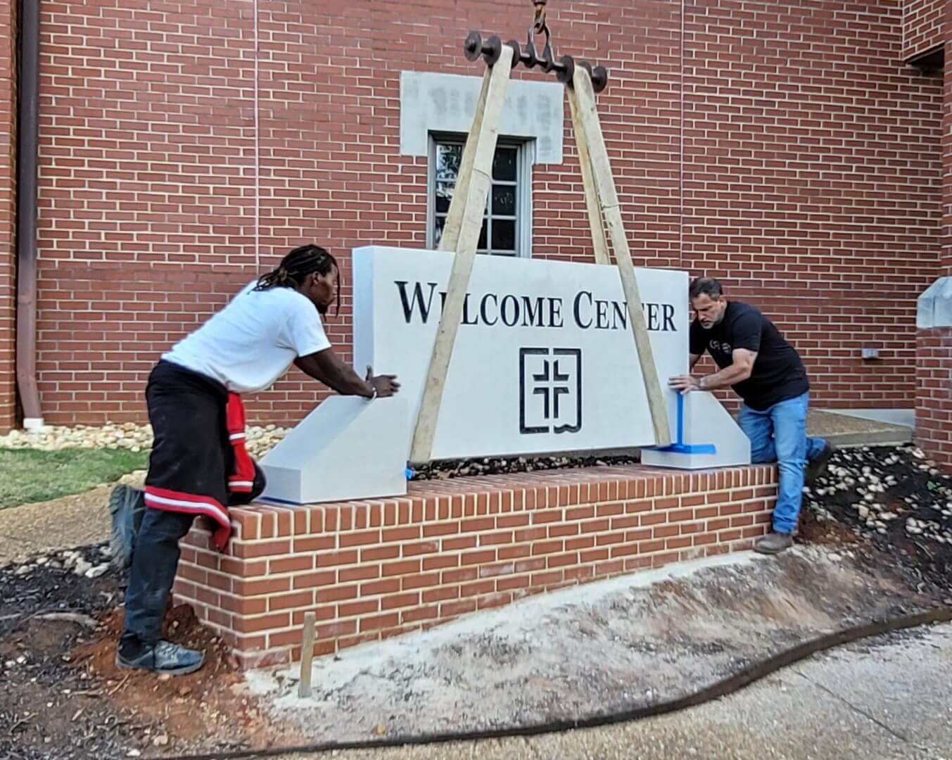 Two men installing a carved stone welcome center sign on a brick base