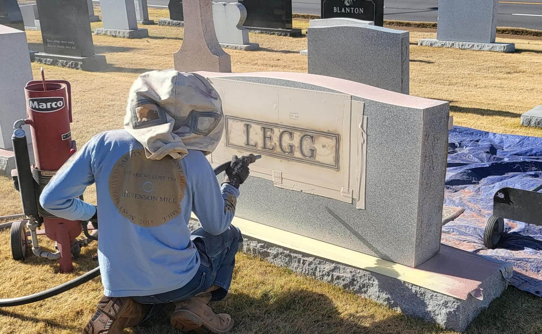 A craftsman in a blasting hood sandblasting a family name into a rose granite monument in a cemetery