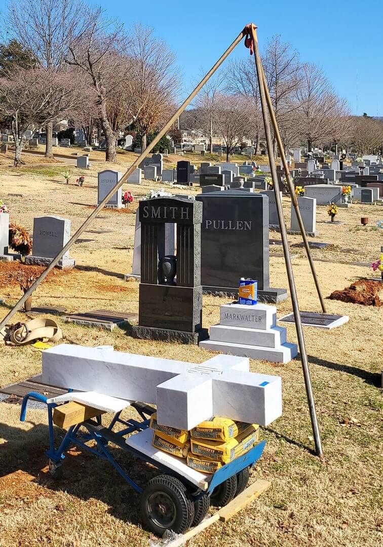 A tripod hoist standing over a pair of upright monuments in a winter cemetery, base sections waiting on a cart
